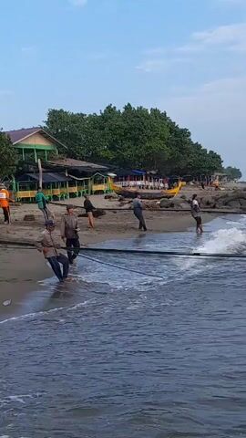 The scene is a beach setting, with a group of people engaged in a fishing activity. There are approximately eight people visible, all standing on the sandy beach near the water's edge. They are pulling on a thick, dark rope that stretches out into the water, presumably connected to a fishing net. The act of pulling suggests they are in the process of hauling the net out of the sea. 

The environment includes a building structure in the background, characterized by a dark-colored roof and light-colored walls, possibly a restaurant or beachside establishment. Trees are visible, creating a natural backdrop. Several fishing boats are also beached nearby, indicating the locale is likely a fishing village or coastal area. The water is blue with white foamy waves gently crashing on the shore. The sky is clear, with sunlight enhancing the overall scene.