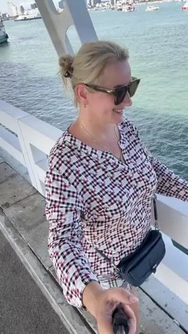 A woman with blonde hair pulled back in a bun, wearing sunglasses, a plaid shirt, and plaid shorts, stands on a white structure, possibly a bridge or pier, holding a selfie stick. She is smiling at the camera. Behind her, the ocean is visible, with boats and a cityscape in the distance under a cloudy sky. She has a dark blue fanny pack slung across her body.