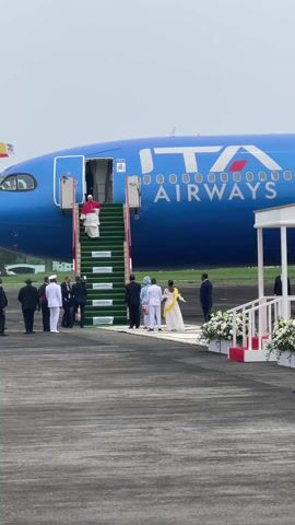 The video captures Pope Francis descending from an ITA Airways airplane. He is dressed in his characteristic white robes with a red cape. A red carpet leads from the foot of the plane's stairs to a small white gazebo adorned with white flowers.  Several people stand at the base of the stairs, likely awaiting the Pope's arrival. These include individuals in suits, white uniforms, and some wearing traditional African attire. The ITA Airways plane is predominantly blue with white lettering. The overall scene suggests an official arrival ceremony.