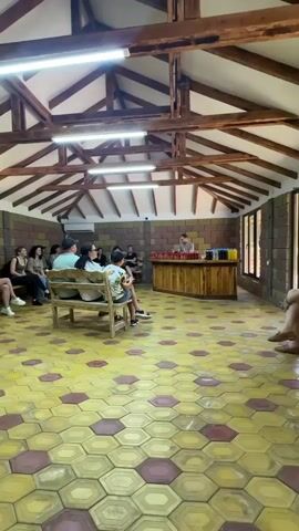 A group of people are gathered in a room with a hexagonal tiled floor. The room has a wooden beamed ceiling with fluorescent lighting. On the left side of the room, several people are sitting on benches, facing a wooden counter at the front. A person stands behind the counter, appearing to be giving a presentation or demonstration. There are bottles or jars on the counter. The walls are made of stone or brick. The overall atmosphere appears to be educational or a tasting event.