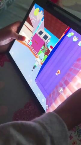 A person is playing a game on a tablet. The game appears to be a simulation or role-playing game with cartoonish graphics. The person is holding the tablet with both hands and their fingers are interacting with the touchscreen. The game screen shows a colorful, isometric view of a virtual environment with buildings and characters. There are also various icons and menus on the screen, suggesting different game functions and options. The lighting in the room is dim, and the tablet screen is illuminated, casting a glow on the person's hands and face. The background shows a bed with a floral pattern.
