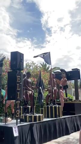 The video captures an outdoor event on a bright, cloudy day. A group of men, mostly shirtless and wearing white briefs or similar attire, are positioned on a raised platform covered with black fabric. The platform is decorated with black and gold striped cylindrical pedestals. Some of the men are holding black flags on poles, and one is waving a black flag. One man on the far right wears a headpiece with feathers. To the left of the platform, a tall black speaker tower with the word "ELITE" in vertical gold lettering is visible, along with a white card displaying the number "5". Behind the platform, there are green trees and palm trees, and in the background, a sign with "ELITE" is present. The overall atmosphere appears to be a performance or parade.