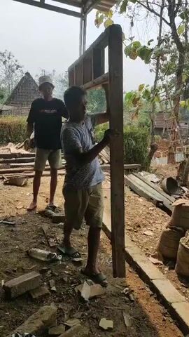 The video shows two young men working on a wooden structure outdoors. The man in the foreground, wearing a striped t-shirt and shorts, is holding a large wooden frame upright, appearing to be installing it. His body is angled as he braces the frame with his hands and body. The second man, standing to the left and slightly behind, wears a black t-shirt, shorts, and a cap. He is also interacting with the wooden structure, his hands visible and his body leaning towards the frame, possibly assisting in its placement or stabilization. They are in an outdoor area with a dirt ground covered in debris, including broken bricks and a plastic bottle. Wooden planks and other building materials are scattered around. In the background, there are trees, a roof, and what appears to be a house with a red and white flag. The overall scene suggests a construction or renovation project taking place in a rural or semi-rural environment.