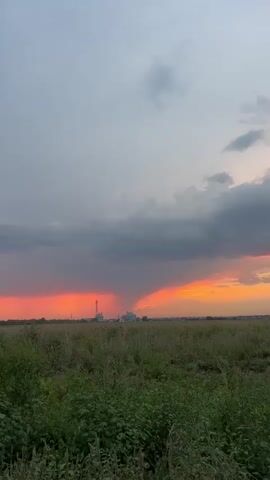 The video shows a wide landscape view during sunset. A field of green grass and plants occupies the foreground. In the middle ground, a distant industrial area with buildings and a tall tower is visible. The sky is filled with dramatic clouds, with a patch of vibrant orange and red sunset light illuminating the scene, partially obscured by dark storm clouds. There are no people or animals visible in the video. The overall impression is one of a stormy sunset over a rural or semi-industrial landscape.