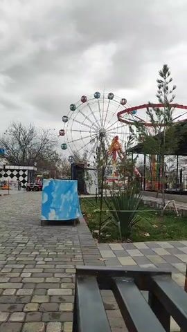 The video depicts a deserted amusement park on an overcast day. The park features a Ferris wheel with multicolored passenger cars, a spinning ride, and several other smaller rides and attractions, all of which appear to be closed. The ground is paved with gray brick and interspersed with patches of grass and young trees. In the foreground, there is a blue box covered with a cloud-printed tarp and a partially visible metal bench. The video is captured from a stationary perspective, providing a full view of the park's layout and vacant ambiance.