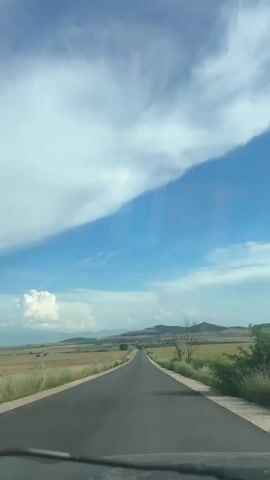 The video shows a view from inside a vehicle driving down a straight, paved road. The road is bordered by fields of dry grass or wheat on either side. In the distance, there are rolling hills and mountains. The sky is blue with large, white clouds, including a prominent band of clouds that stretches across the frame. A bare tree is visible on the right side of the road. The perspective suggests the video is captured from the driver's or front passenger's seat.