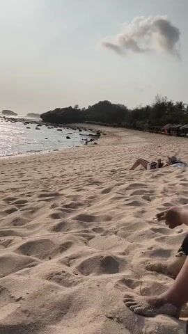 The video shows a sandy beach scene with several people. In the foreground, a person's legs and feet are visible, with their right hand resting on the sand. To the left and further down the beach, another person is lying on their stomach, facing away from the camera, wearing a hat and shorts. They appear to be reading or looking at a phone. The beach stretches into the distance, with a line of trees and small huts visible along the shoreline. The ocean is to the left of the frame, with gentle waves lapping the shore and some rocky outcrops in the distance. The sky is bright with a few scattered clouds. The overall impression is a relaxed beach setting.