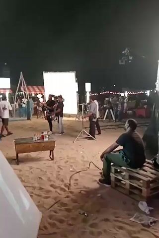A nighttime shot captures a film set in a sandy, outdoor location. A large, rectangular white reflector stands prominently on the left. In front of it, several crew members are actively working, including one operating a camera. Further back, a camera is mounted on a dolly track. On the right, another crew member sits on a wooden pallet, observing the scene. String lights crisscross in the background, illuminating distant structures.