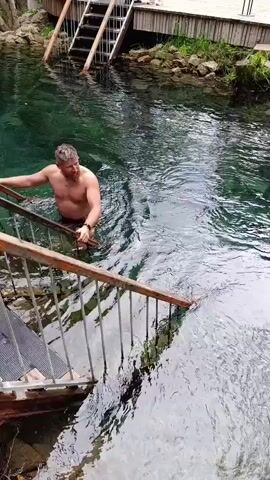 A shirtless man with short, brown hair and a beard swims in a clear, greenish body of water. He is holding onto the railing of a set of wooden stairs with metal supports, which descend into the water. The man is facing the camera and appears to be moving towards the stairs. In the background, a wooden dock or deck is visible with another set of stairs leading down to the water. The water is calm, with ripples emanating from the man's movements. The surrounding area appears to be natural, with rocks and greenery visible along the edge of the water. The overall scene suggests a recreational activity in a natural setting, possibly a lake or a spring. There is no visible text in the media file. The lighting suggests it is daytime, and the weather appears to be pleasant.