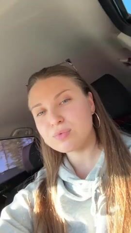 A woman in a light gray hoodie is sitting in the driver's seat of a car. She has long, straight, brown hair and blue eyes. She is wearing gold hoop earrings and sunglasses are perched on top of her head. The background of the video is the interior of the car. Music plays. The woman smiles.