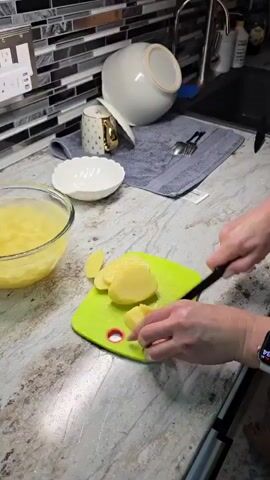 A close-up shot shows a person's hands slicing potatoes on a green cutting board. The cutting board is on a granite countertop. To the left of the cutting board is a glass bowl filled with sliced potatoes. Behind the cutting board, there is a sink area with a dish towel, a white bowl, and some cutlery. The background features a tiled backsplash. The person is wearing a smartwatch.