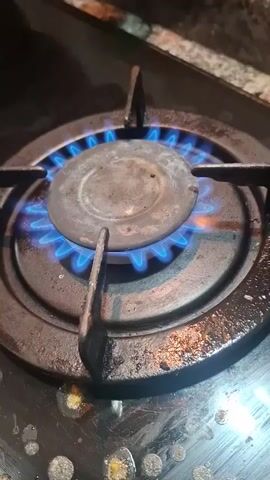 The video shows a close-up of a lit gas stove burner. The burner is circular and made of metal. The flame is blue and evenly distributed around the burner. The stove top around the burner appears to be dirty with some oily residue and speckles of debris. The metal grate above the burner is also visible.