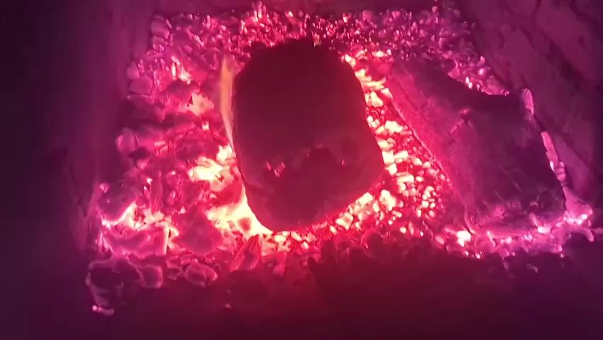 The scene is inside a brick fireplace, glowing with a vibrant pink and red light from burning embers and small flames. A partially charred log lies on the right side, while a larger, wrapped object rests in the center of the glowing embers. The fireplace's interior is dark and indistinct around the bright, burning area.