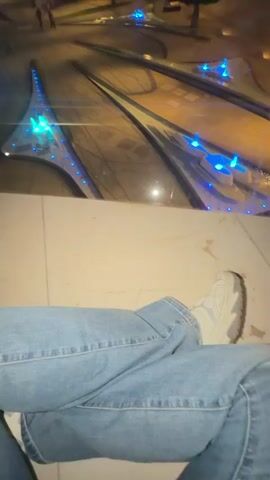 The video shows a low-angle view looking down at a modern, geometric plaza at night. The plaza features sleek, curved structures illuminated by vibrant blue lights, resembling water features or artistic installations. The ground is paved with stone. In the foreground, the legs of a person wearing blue jeans and white sneakers are visible, sitting with their legs crossed. Further in the background, a few individuals can be seen walking or standing in the plaza, and trees are present. The overall atmosphere is urban and contemporary, with the artificial lighting creating a dramatic effect in the darkness.
