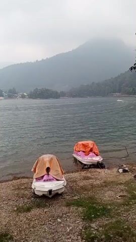 The video shows a wide shot of a lake on a cloudy day. In the foreground, on the rocky shore, are two jet skis covered with tarps. One jet ski is mostly white with a pink and orange tarp. The other is white with an orange tarp. There are some tires scattered on the shore as well. In the background, across the lake, are trees and some buildings. Hills or mountains are visible in the distance, shrouded in mist. The water is choppy, and there is some movement on the water in the distance, possibly other boats. The overall atmosphere is peaceful and overcast.