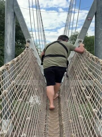 A person wearing a harness and olive green t-shirt walks across a rope bridge with netting on either side. They are barefoot and wearing black shorts. The bridge has a rope and sand path in the center. A sign at the top of the bridge reads "Hängebrücke" and "Barfußpfad" in German. The background is green trees and a cloudy blue sky.