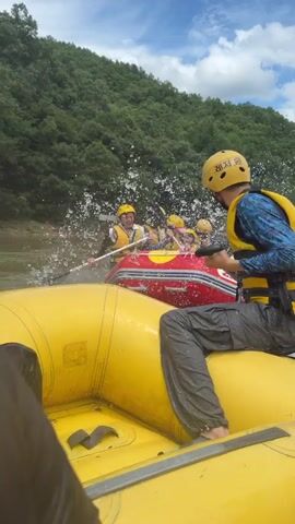 A group of people are whitewater rafting. There are at least two rafts visible. One raft is yellow and in the foreground. The second raft is red and white and further back in the water. Both rafts appear to be in motion. People are wearing yellow helmets and yellow life vests. One person in the foreground raft is wearing a blue long-sleeved shirt and dark pants, and is holding a paddle. This person is looking to the left. In the red and white raft, there are multiple people holding paddles and facing forward. They are in a river surrounded by lush green trees and a blue sky with white clouds. Water is splashing in the air, indicating the rafts are moving through choppy water.