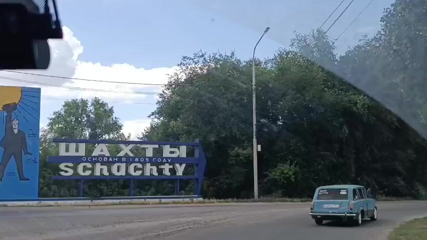 The video shows a view from inside a vehicle, looking out the front windshield and side window. A blue sign with white lettering is visible on the left side of the road, with the words "ШАХТЫ" and "SCHACHTY" written on it. Below these words, smaller text reads "ОСНОВАН В 1805 ГОДУ". The sign has an arrow pointing to the right. Dense green trees and foliage line the right side of the road. A light-colored utility pole with a lamp is also present. A light blue car is driving away from the camera on the road to the right. Another darker car is visible further down the road. The sky is visible above the trees and appears to be cloudy. A black object, possibly a dashcam, is mounted in the upper left corner of the windshield. The road surface appears to be asphalt. The overall scene suggests the vehicle is traveling on a road leading into or out of a town named Shakhty.