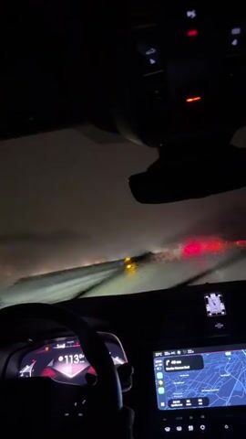 The video shows the view from inside a car driving at night on a highway. The road is wet, likely from rain, and the windshield wipers are in use. There are multiple red taillights of other vehicles visible ahead, indicating traffic congestion. The car's dashboard displays a speed of 112 and a navigation map. The overall environment is dark, with the only significant light sources being the car's interior lights, headlights from other vehicles, and the illuminated dashboard. The driver's hands are visible on the steering wheel. The video appears to capture a moment of challenging driving conditions due to weather and traffic.