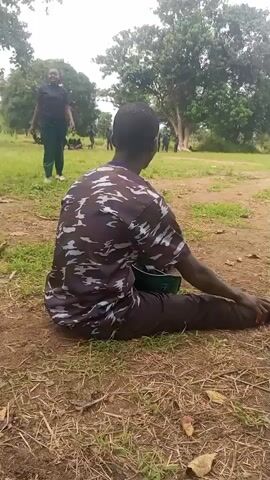 A person in a camouflage shirt and dark pants sits on the ground, stretching their leg. Another person in dark clothing walks in the background, and several other people are visible further in the distance. The scene appears to be outdoors, with trees and grass in the environment. The sky is overcast.
