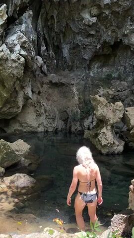 A woman with blonde hair wearing a black bikini enters a body of water within a cave. She submerges herself in the water and begins to swim. The water is clear and the cave walls are rough and rocky.