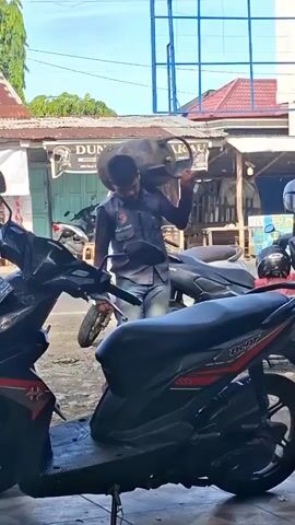 A man is carrying a large, cylindrical object, possibly a gas tank, on his right shoulder. He is wearing a dark blue uniform shirt with a logo on the left chest, blue jeans, and sandals. He appears to be walking in a street or alleyway. Several motorcycles are parked in the background. The environment looks like a commercial area with buildings and signage. The lighting suggests it is daytime.