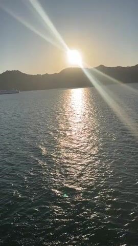 The video shows a time-lapse of a sunset over a body of water. The sun is setting behind a silhouetted mountain range. The sun's rays are casting a bright reflection on the water. A ship is visible in the distance on the left side of the frame. The sky transitions from a light blue to a purple hue as the sun sets.
