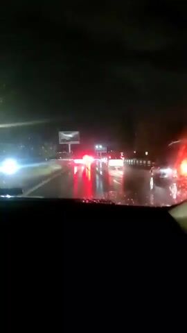 The video shows a view from inside a vehicle driving at night on a wet road. Rain is visible hitting the windshield, creating streaks of water. The road ahead is lit by the headlights of other vehicles, which appear as bright white and red lights reflecting off the wet surface. A billboard is visible on the left side of the road. The overall scene is dark and the conditions suggest poor visibility due to the rain and darkness.