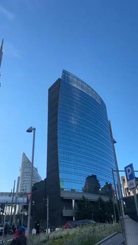 The video shows a low-angle view of modern skyscrapers against a clear blue sky. The primary focus is on a tall, curved skyscraper with a reflective blue glass facade. To the left, another skyscraper with a similar curved design and a pointed spire is visible. In the background, a smaller, triangular-shaped building can be seen. The street level shows a pedestrian walkway, some people walking, and cars on the road. There are also streetlights and some greenery. The video appears to be a panning shot, moving slightly to reveal different angles of the buildings. The lighting suggests it is daytime.