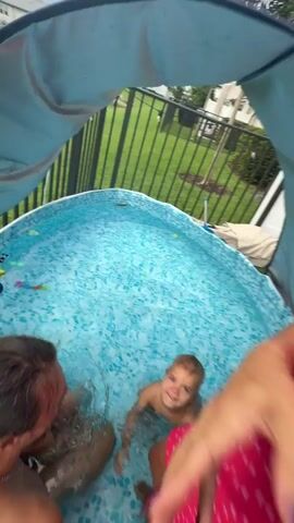 A family is having fun in a small inflatable swimming pool set up in a backyard. The pool is filled with clear blue water and has a patterned interior. Toys are visible floating in the pool, including what appear to be colorful balls and perhaps a toy boat. A black metal fence encloses the yard, with green grass and some trees visible beyond it. A light-colored canopy extends over part of the pool, providing shade.

A man with long hair pulled back is in the pool, facing a young child. He is smiling and appears to be interacting with the child. The child, who is blonde and appears to be a toddler, is in the water and smiling broadly.

A woman, wearing a red dress with a white heart pattern, is also in the pool. She is interacting with the child and appears to be laughing. Her upper body is visible, and she is holding the child.

The perspective of the video is from someone looking over the edge of the pool into the water. In some shots, a person's legs and feet are visible in the foreground, partially submerged in the water. The overall atmosphere is one of joyful family recreation.