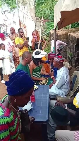 Here is a description of the video:
The video shows a group of people gathered in what appears to be a casual outdoor setting, possibly a backyard. A table is set up with a box of "LORD'S" beverage, some fruit, and other items. Several individuals are seated around the table, engaging in conversation. One man, wearing a red and black patterned hat, is speaking into a microphone. Other people are standing around the table, some recording or taking pictures with their phones. The background features greenery, a wall, and other structures. The overall atmosphere suggests a social gathering or event, with an informal and celebratory tone.