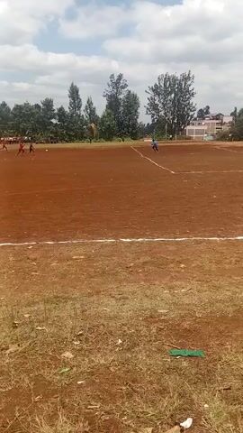 The video depicts a soccer game being played on a dirt field with white boundary lines under a partly cloudy sky. Several players, mainly wearing red jerseys, are scattered across the field, actively engaged in the game. Trees line the perimeter of the field, and a building is visible in the background. The foreground shows grassy areas with some scattered debris. A man wearing black shoes, a green shirt and carrying a dog or small animal runs across the bottom right corner of the video.