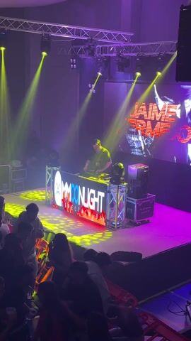 The video shows a DJ performing on a stage at what appears to be a club or event. The DJ is standing behind a booth with the word "MOONLIGHT ESPECTACULOS" illuminated on the front. Behind him is a large screen displaying various graphics. The stage is lit with multicolored lights. A crowd of people stands in front of the stage, watching the performance. Some people are holding phones and appear to be recording the event. The overall atmosphere is that of a lively, energetic party or performance.