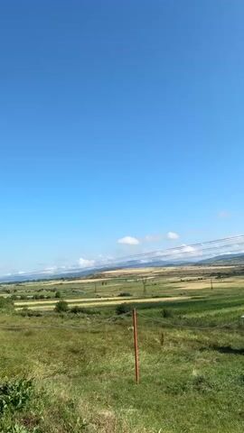 A wide shot captures a rural landscape under a clear blue sky with scattered clouds. In the foreground, tall green grass dominates, with a single red-topped metal post partially visible. To the right, a small, metallic shed with a corrugated roof is situated. The midground reveals a patchwork of agricultural fields, displaying varied shades of green and yellow, suggesting different crops or stages of growth. Two power line towers are prominent features in the distance. Beyond the fields, rolling hills form the horizon, receding into a hazy blue. Overhead, power lines stretch across the frame, adding a man-made element to the natural scenery. The sun is shining, illuminating the landscape.