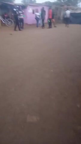 The video shows a blurry view of a dirt area, presumably a street or open space, with several people standing or walking around. There are a few structures visible in the background, including what appears to be a small shack or shop and a pink-colored building. A bicycle is parked near the shack. The video is somewhat shaky and lacks focus, making it difficult to discern fine details. The lighting suggests it is either early morning or late afternoon.