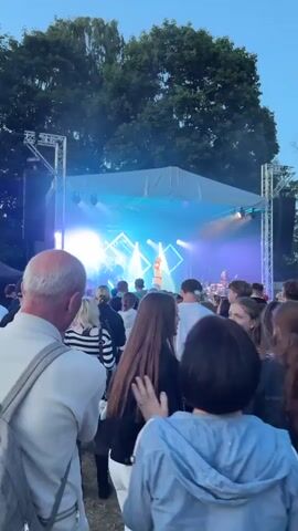 A low-angle vertical video captures an outdoor concert at dusk. A stage with bright blue and white lights is set up in the background, with a performer visible center stage. A crowd of people stands in the foreground, facing the stage. The audience members are diverse in age and gender, with many appearing to be young adults. The atmosphere is lively, with some audience members looking towards the stage and others interacting with each other. Trees are visible behind the stage, and the sky is a dusky blue, indicating it is late evening. The overall impression is that of a vibrant outdoor music event.