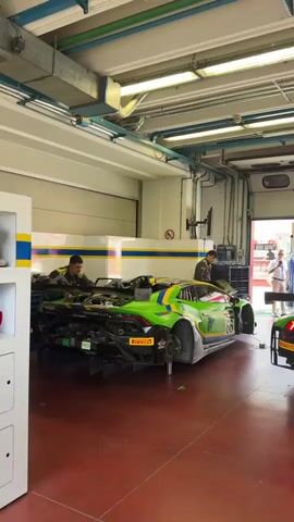 The video shows a group of people working on two green and yellow race cars inside a garage. The cars are Lamborghinis, identifiable by their distinctive body shapes and branding. Several individuals in racing attire are actively engaged in maintenance, with tools and equipment visible around the vehicles. One car is elevated on jacks, with its rear wheels removed, suggesting it is undergoing significant mechanical work. The garage environment is characterized by industrial lighting, exposed pipes and wiring, and a red, rubberized floor. The overall atmosphere appears to be a pit stop or workshop setting within a racing facility.