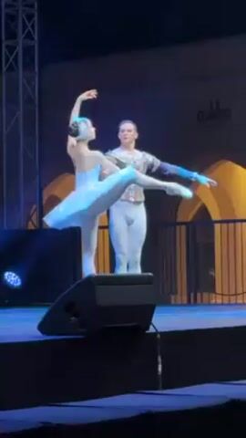 Two ballet dancers, a woman and a man, are performing on a stage. The woman is in a white tutu and tiara, standing en pointe with her leg extended. The man is in a matching outfit, supporting her. They are both illuminated by blue stage lights. A large black speaker is in the foreground, slightly obstructing the view. The background includes a metal tower structure and part of a building with illuminated archways.