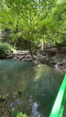 This video captures a tranquil natural scene featuring a clear, turquoise pool of water at the base of a cascading waterfall. The water flows over tiered rock formations, creating a series of small drops. Lush green trees with dense foliage surround the pool, providing a shaded canopy. The sunlight filters through the leaves, creating dappled patterns on the water's surface and the rocks. The environment appears to be a forest or a park with well-maintained wooden pathways and railings visible in the background and foreground, suggesting it is a place accessible to visitors. The overall atmosphere is serene and peaceful.