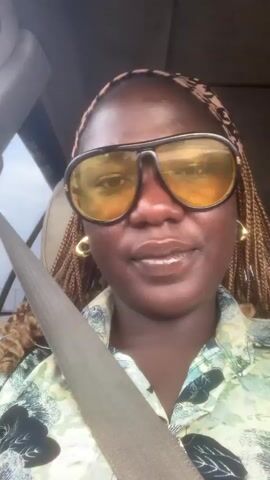 An adult female is filmed in a close-up selfie video inside a vehicle. She has dark skin and is wearing large black-framed sunglasses with yellow-tinted lenses. A light-colored headband with a dark houndstooth pattern is on her head, and her hair is styled in thin, light brown braids. She wears large gold-colored hoop earrings and a light green button-up shirt with a dark abstract or leaf pattern. A gray car seatbelt diagonally crosses her chest. She is smiling, showing her teeth, and looking directly at the camera. The background behind her is blurred, indicating the interior of a car.