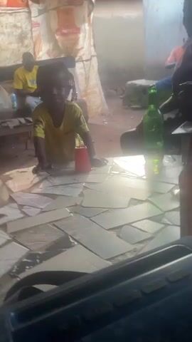 The video shows a young child in a yellow t-shirt standing at a table covered with broken tiles. The child is reaching towards a red cup on the table and appears to be interacting with an unseen person whose hand briefly enters the frame and touches the child's head. In the background, two adults are seated, and there are bags and other objects in a rustic environment. The lighting suggests it is daytime, possibly outdoors or in a poorly lit indoor space. The overall impression is a casual, everyday scene.