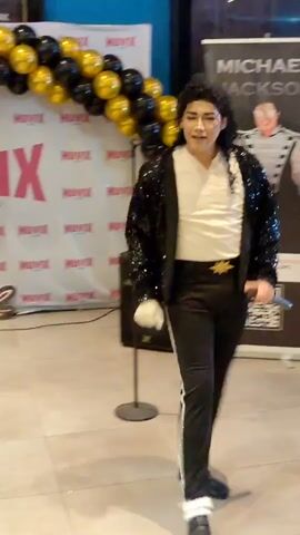 A male impersonator dressed as Michael Jackson, wearing a sequined black jacket, white shirt, black pants with white stripes, and white ankle cuffs, performs. He is standing in front of a banner with Michael Jackson's image and text, and a balloon arch made of black and gold balloons. A speaker is positioned to the left of the banner. Another banner is partially visible to the right. The floor appears to be tiled.
