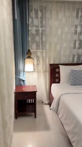 The video features a slow panning shot of a twin bedroom from left to right. The scene opens with a dark wooden bedside table with a small pink object on top, positioned next to a hanging glass and metal lamp. To the right of the bedside table are two single beds with dark wooden headboards and frames. Each bed is made with white bedding, a white pillow, and a smaller blue-patterned decorative pillow. On the bed to the left, a white rolled towel and a small yellow sticky note are visible. The bed to the right also has a white rolled towel. Behind the beds, a window is covered by white sheer curtains with a patterned design, overlaid by solid light blue curtains pulled to the sides. As the camera pans further right, a large woven, fish-shaped wall decoration is seen mounted above the right bed, with a white air conditioning unit installed above it on the wall.