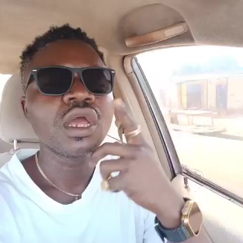 The video shows a Black man wearing a white t-shirt, black sunglasses, a gold chain, a gold watch, and a gold ring, in the driver's seat of a car. He speaks while looking directly at the camera. The car appears to be in motion. A building is visible through the car window.