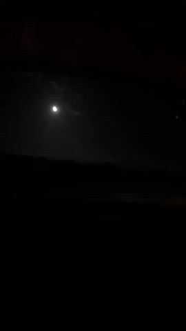 The video begins with a view of a dark night sky with a bright full moon. The camera then pans to the interior of a car, showing the dashboard and steering wheel illuminated by red lights. The scene shifts to a view from the car's windshield, looking out at a dark, empty road. The headlights of the car illuminate the road ahead, which is lined with white markers. The road appears to be winding and leads into the darkness. The video ends with a shot of the road stretching into the night.