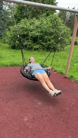A young woman with blonde hair, wearing a gray hoodie and blue shorts, is swinging on a tire swing in a park. She is lying on her back on the tire, her legs extended, and her arms holding onto the chains of the swing. The swing is attached to a metal frame, with a wooden post visible to the right. The ground beneath the swing is covered in red rubber mulch, and green grass and trees are visible in the background. The video appears to be a short clip of the woman enjoying the swing.
