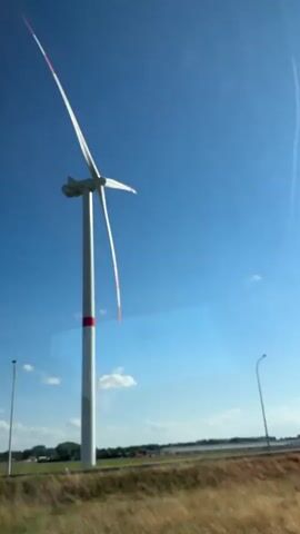 The video shows a tall white wind turbine with red bands on its tower under a bright blue sky with some wispy clouds. The turbine's blades are positioned at an angle. In the foreground, there is a grassy area with some dry grass and a concrete barrier. A lamppost is visible to the left of the wind turbine, and a road is present in the middle ground. In the background, there are fields and some buildings. The sun is shining brightly, creating a lens flare effect in the upper right portion of the frame. The camera appears to be moving, suggesting the video was shot from a vehicle.