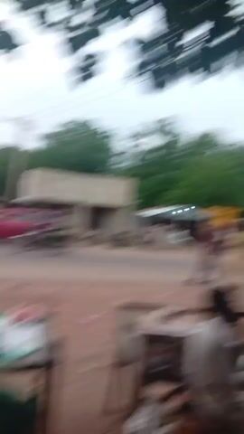 The video begins with a blurry image of trees against a cloudy sky. The scene then shifts to a street scene with people, vehicles, and buildings. In the foreground, two men are standing near a wooden stall. One man, wearing a white shirt, is looking down at something in his hands. The other man, also in a white shirt, is facing away from the camera. Several motorcycles are parked in the background. The street is dusty and appears to be unpaved. In the distance, there are several buildings, including a small shop with a yellow sign. A red car is driving away from the camera. The sky is overcast, and the overall lighting suggests it is either early morning or late afternoon. The video ends with a shot of trees against a hazy sky.