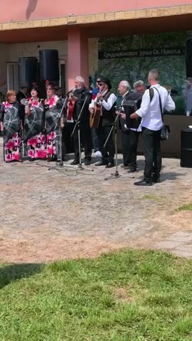 A musical group consisting of both men and women performs outdoors in front of a red and white building with a sign above the entrance in a language other than English. The sign includes an image of trees. The group consists of three women wearing the same patterned long dresses with flowers and four men. One man plays guitar, one plays accordion, and the others sing into microphones. A cameraman stands on the right side of the group facing them and pointing his camera at them. There is a Bulgarian flag on the building. The group stands on a concrete area beside a grassy lawn.
