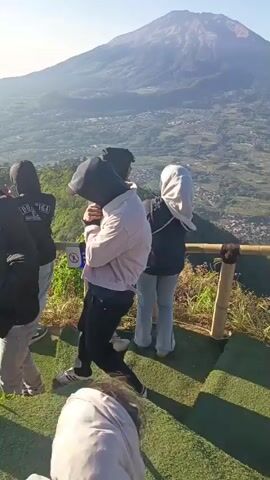 A group of young people are standing on a grassy, elevated platform overlooking a vast valley. Some are wearing hoodies and jackets, suggesting a cooler climate or higher altitude. One person is holding up a phone to take a picture or video of the scenery. The landscape is characterized by rolling hills covered in lush green vegetation, with a town or village visible in the distance. The sun is shining brightly, casting long shadows. The overall impression is one of a scenic overlook where a group of friends are enjoying the view.